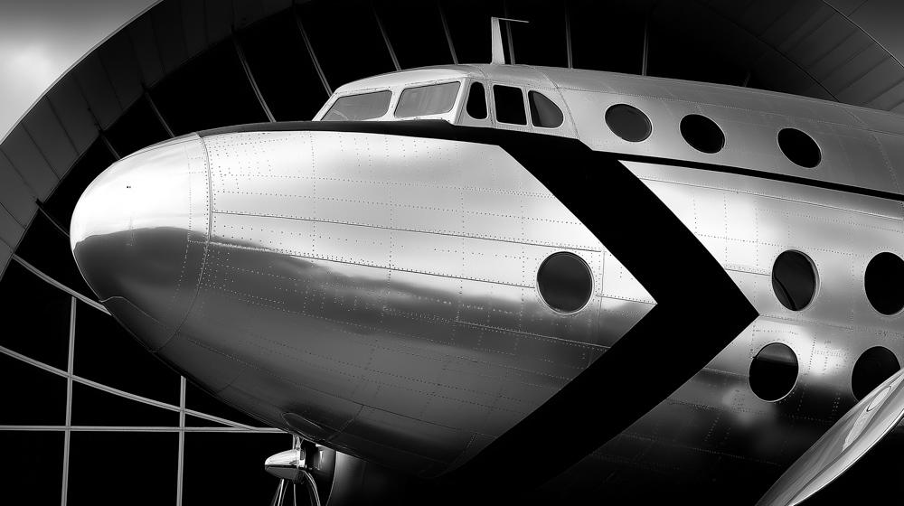 Close-up black and white photo of a vintage aircraft's nose and fuselage with distinctive round porthole windows, displayed in a hangar.