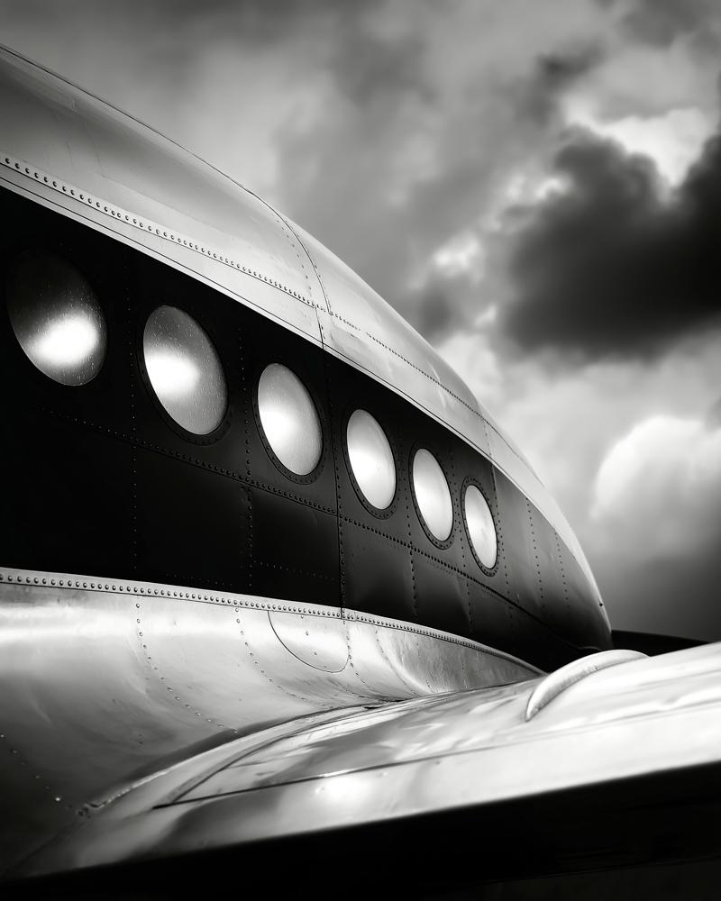 Close-up black and white photograph of a vintage airplane fuselage showing a row of circular windows along its metallic body against a cloudy sky.