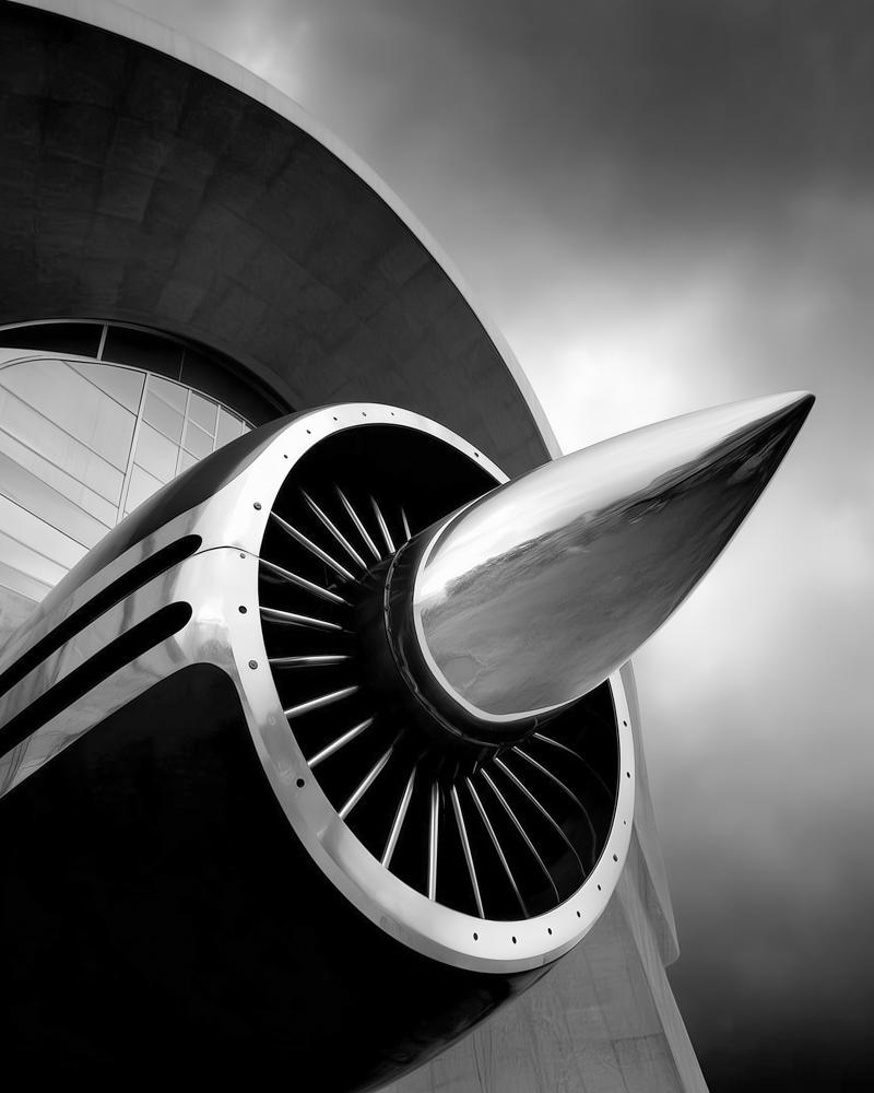 Close-up black and white photo of an airplane jet engine and propeller blade against the curved fuselage of an aircraft.