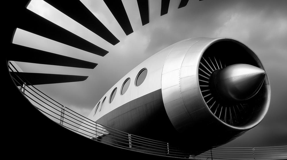 Black and white photograph of an airplane engine and fuselage shot from below, with dramatic angular architectural beams visible in the upper left corner.